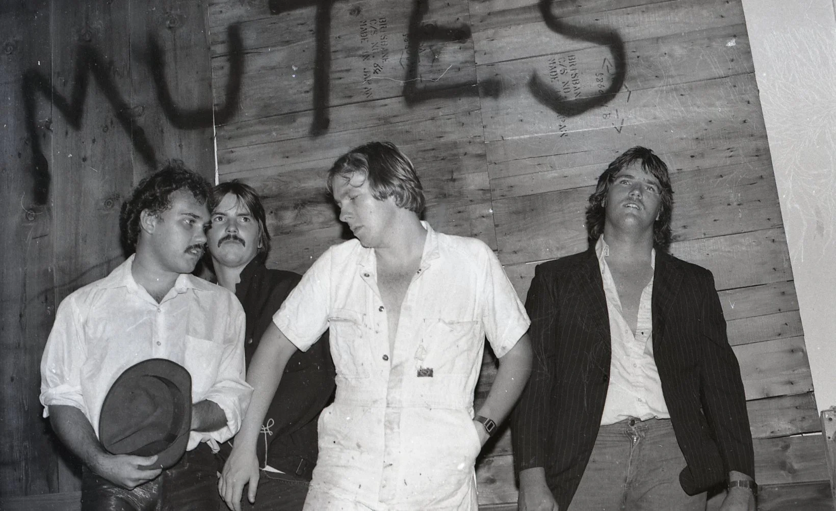 Black and white photo of four men standing against a wooden wall, engaged in a conversation. One man is holding a hat, another is wearing a white shirt with rolled-up sleeves, and the others are dressed in darker clothing, one with a pinstripe jacket