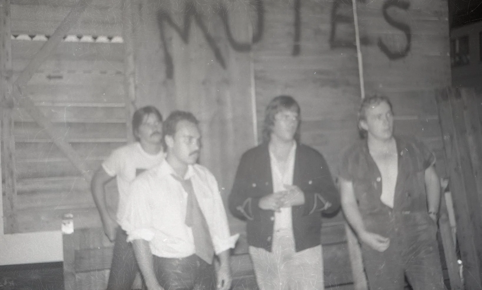 Four men standing indoors in front of a wooden wall, with a man in the background carrying a child on his shoulder. One man is holding something in his hands; the others are standing with their hands by their sides. The photo appears to be old and bl
