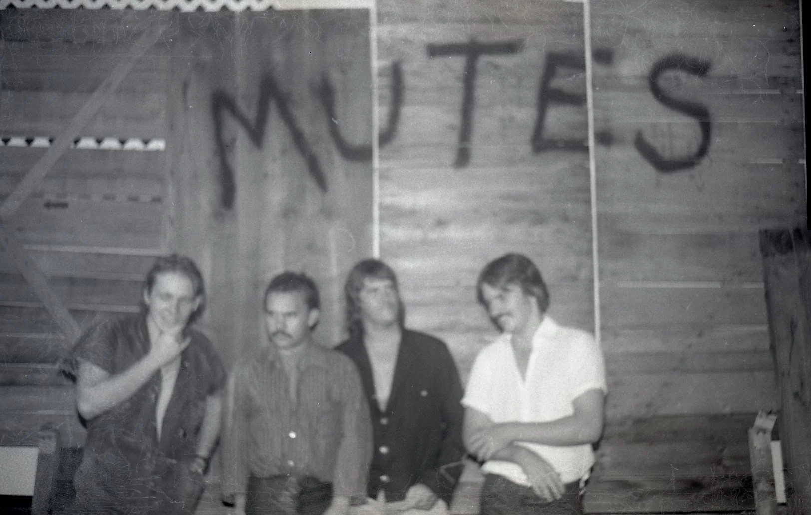 A black-and-white photo of four people standing indoors in front of a wooden wall with the word 'MUTES' written on it. The group consists of two men and two women, dressed in vintage clothing.
