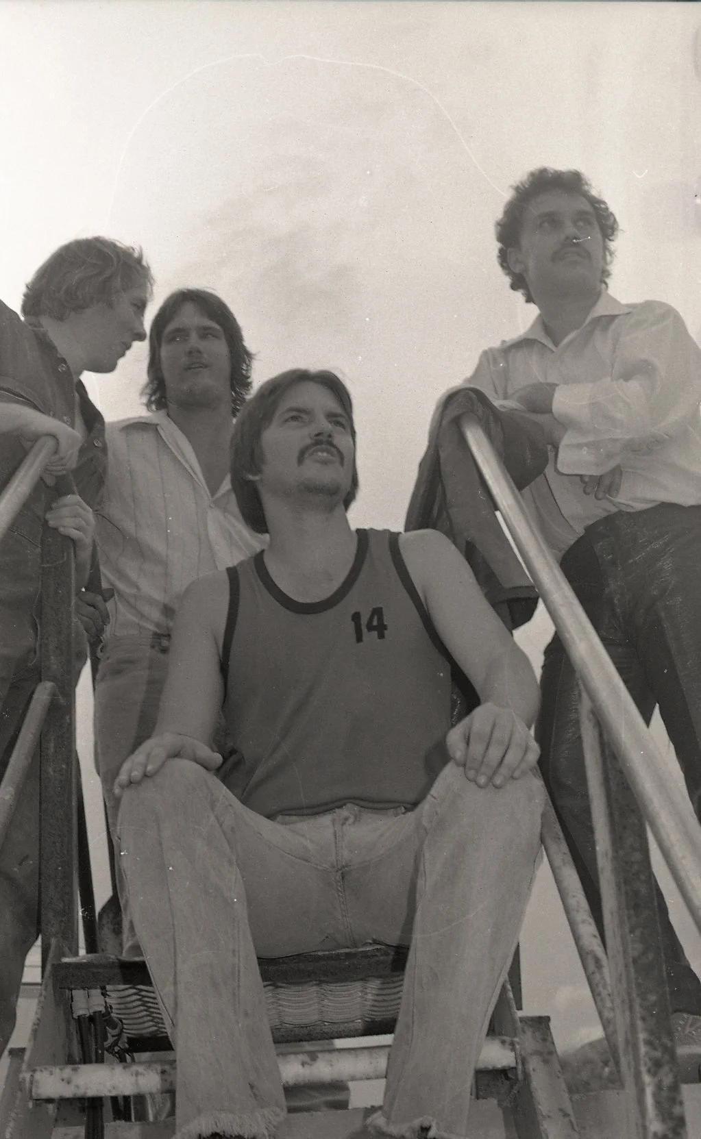 Four men, one wearing a sleeveless shirt with the number 14, sit on outdoor stairs with metal railings, while two others stand nearby. The photo is in black and white and appears to be from the 1970s or 1980s.