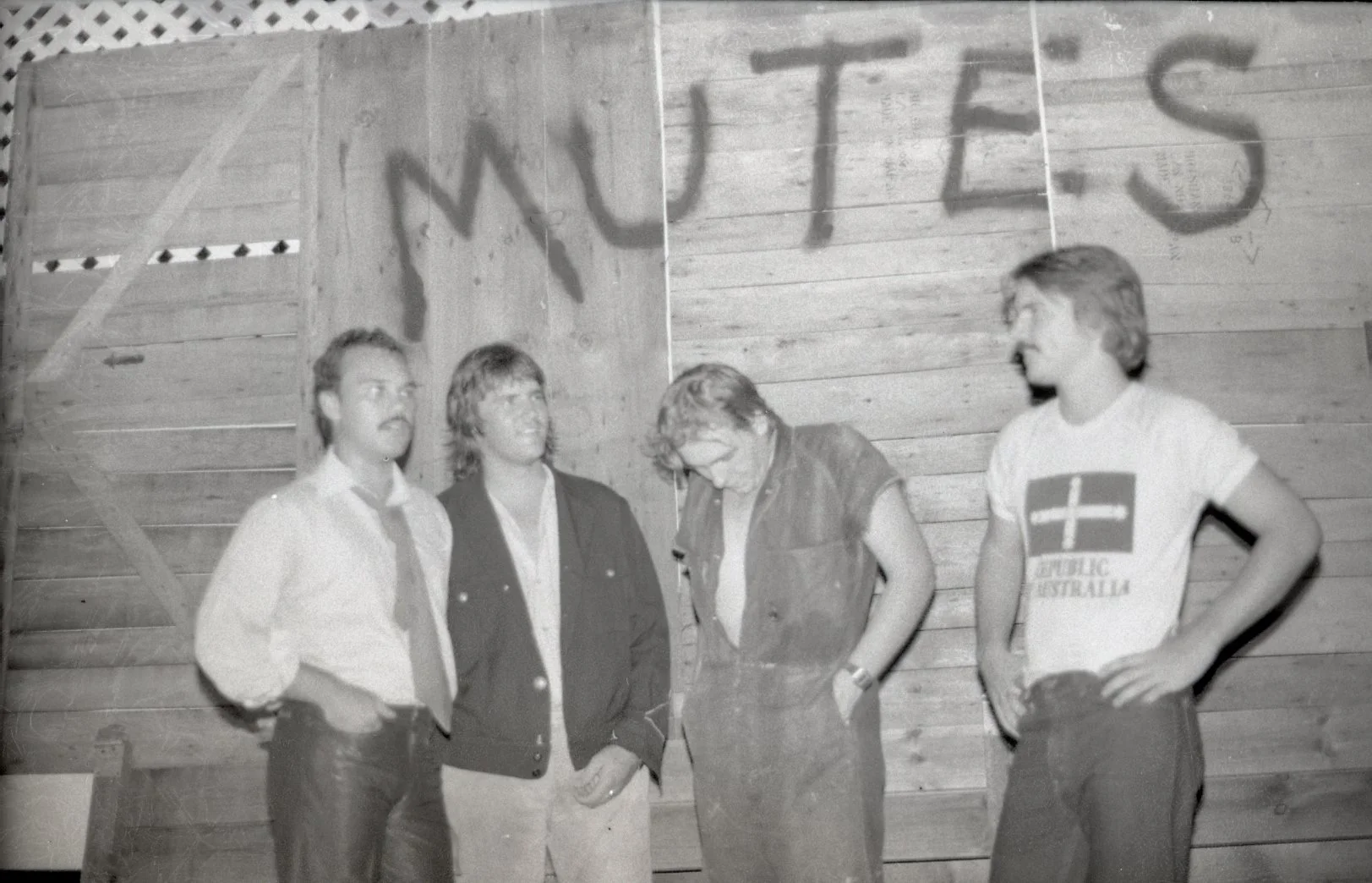 Four men stand in front of a wooden wall with graffiti that reads 'MY LITS'.
