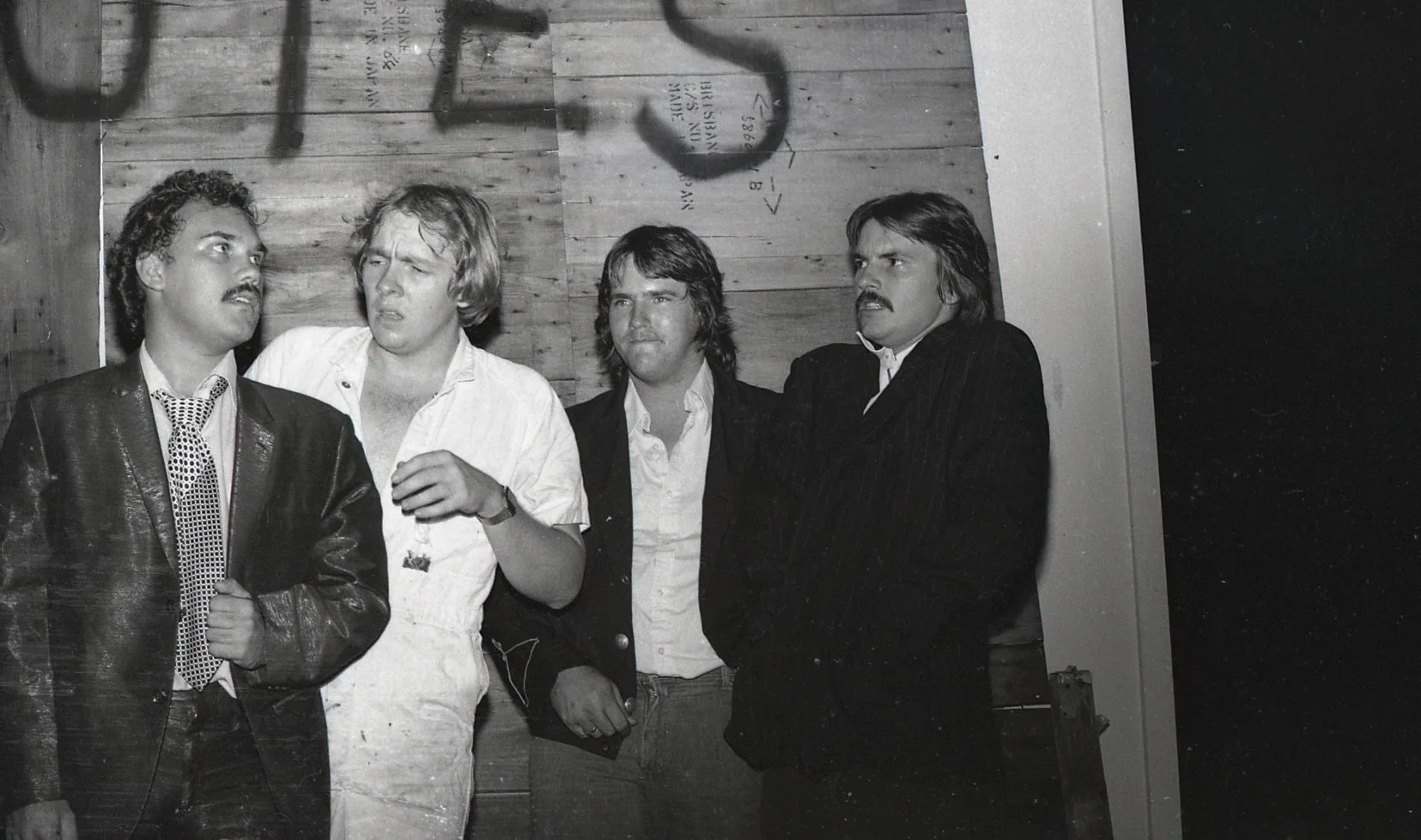 Black and white photo of four men standing together, some showing expressive faces or gestures, in a room with wooden walls.
