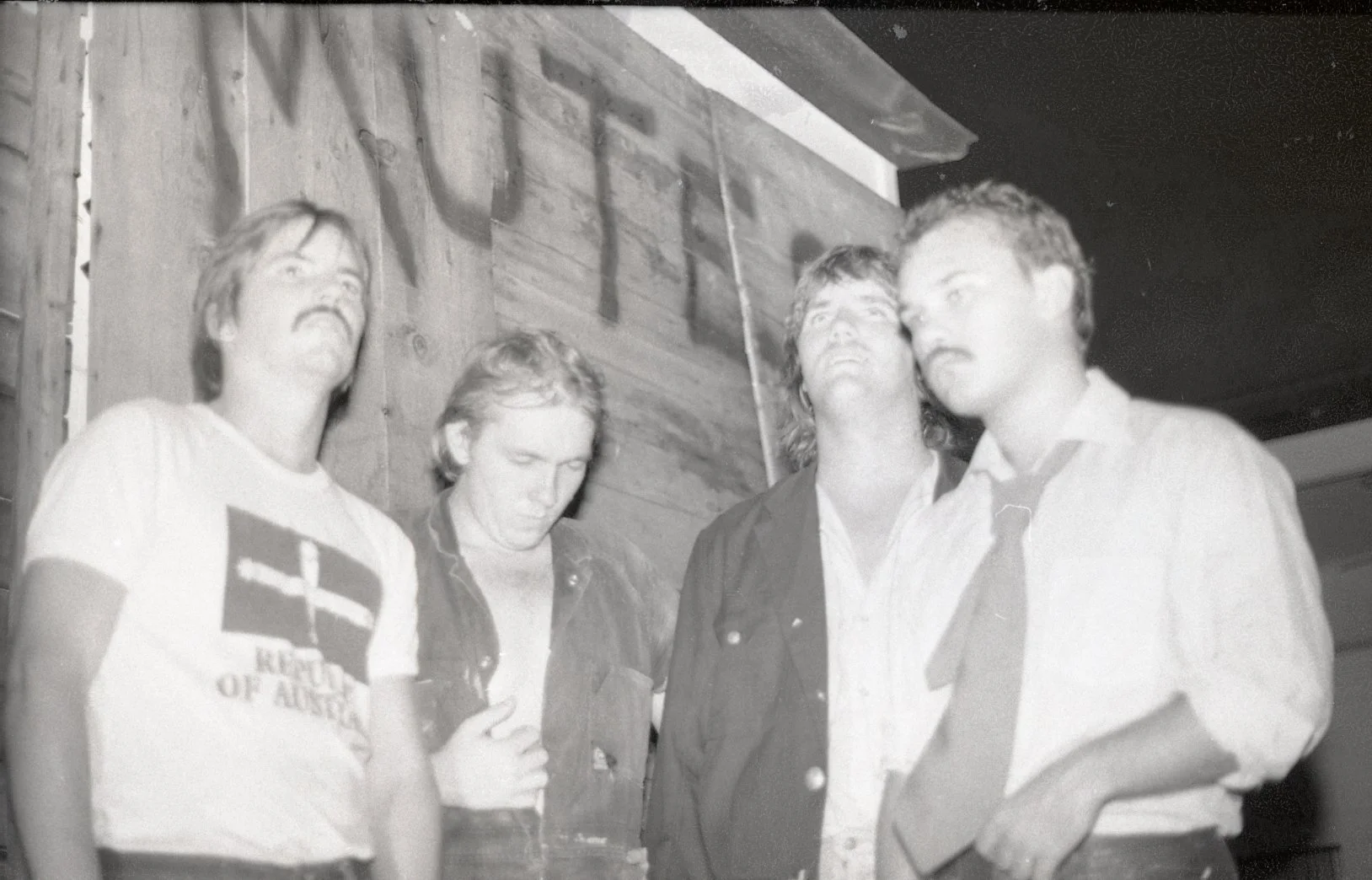 Four young men standing together, two of them with eyes closed, one looking downward, and one staring into distance, in a casual indoor setting with wooden wall behind them and a partially visible sign reading 'HUT'.