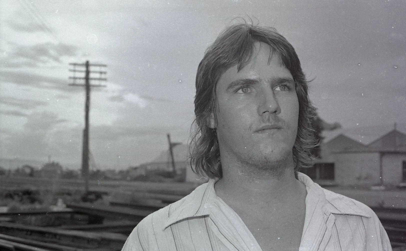 Black and white photo of a man with medium-length hair, looking into the distance. The background shows electrical utility poles and a few houses in a rural landscape.
