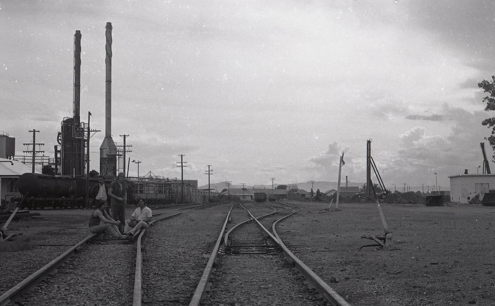 Black and white photo of a railroad yard with train tracks extending into the distance. Five people are sitting and standing near the tracks, with industrial buildings and equipment, such as cranes, tanks, and power lines, visible in the background a