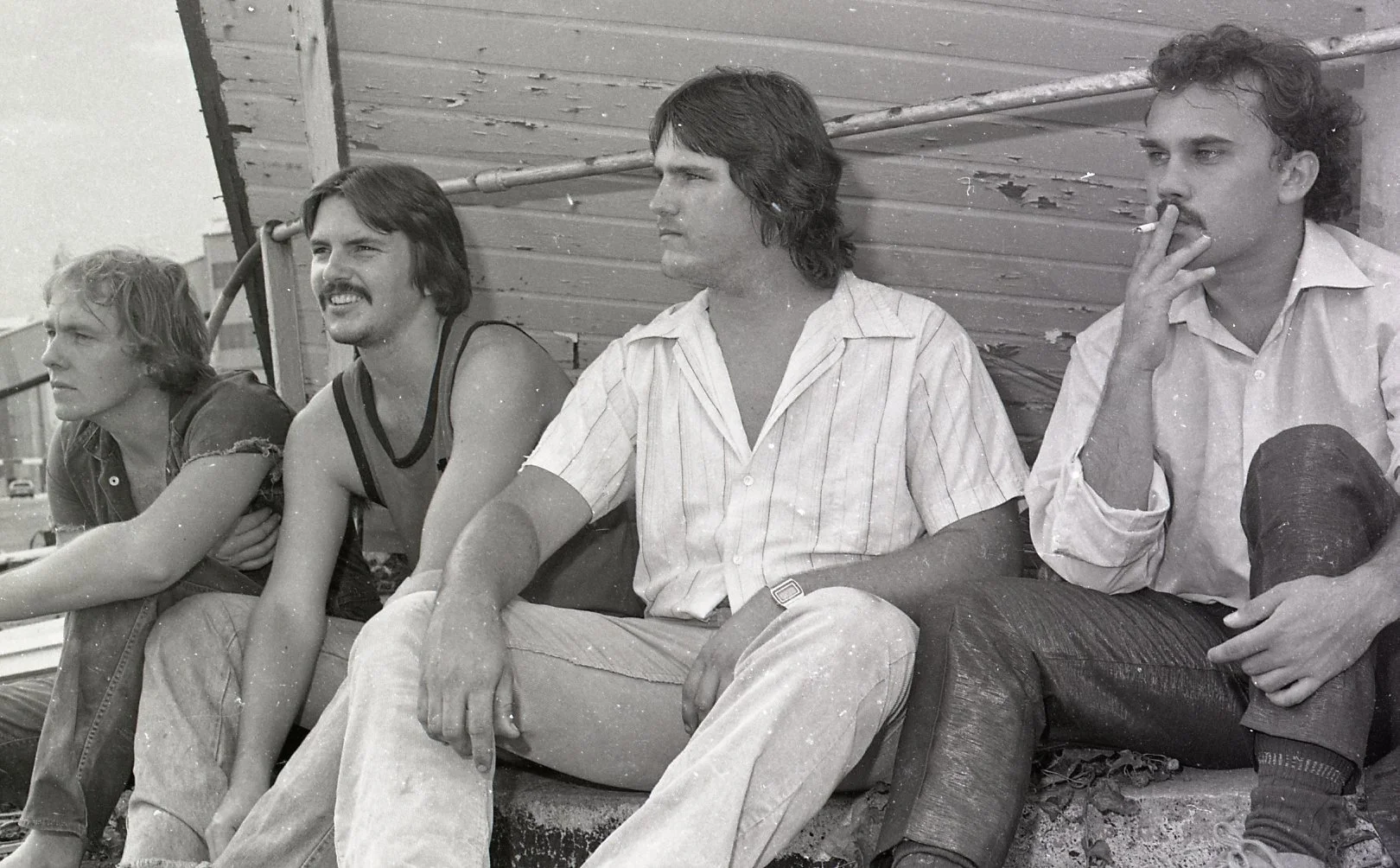 Four men sitting on a bench next to a wooden wall, with one smoking a cigarette; black and white photo.