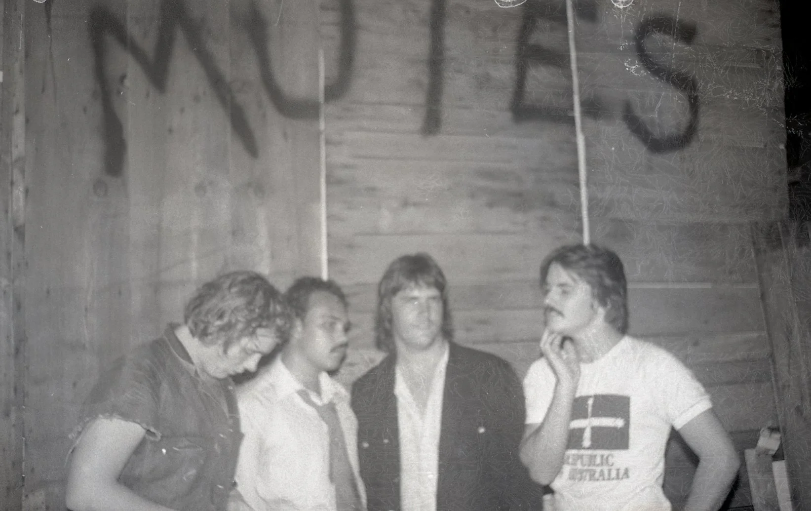 Four people standing indoors in front of a wooden wall, engaged in conversation. One person has a shirt with an Australian flag and the words 'REPUBLIC AUSTRALIA'.