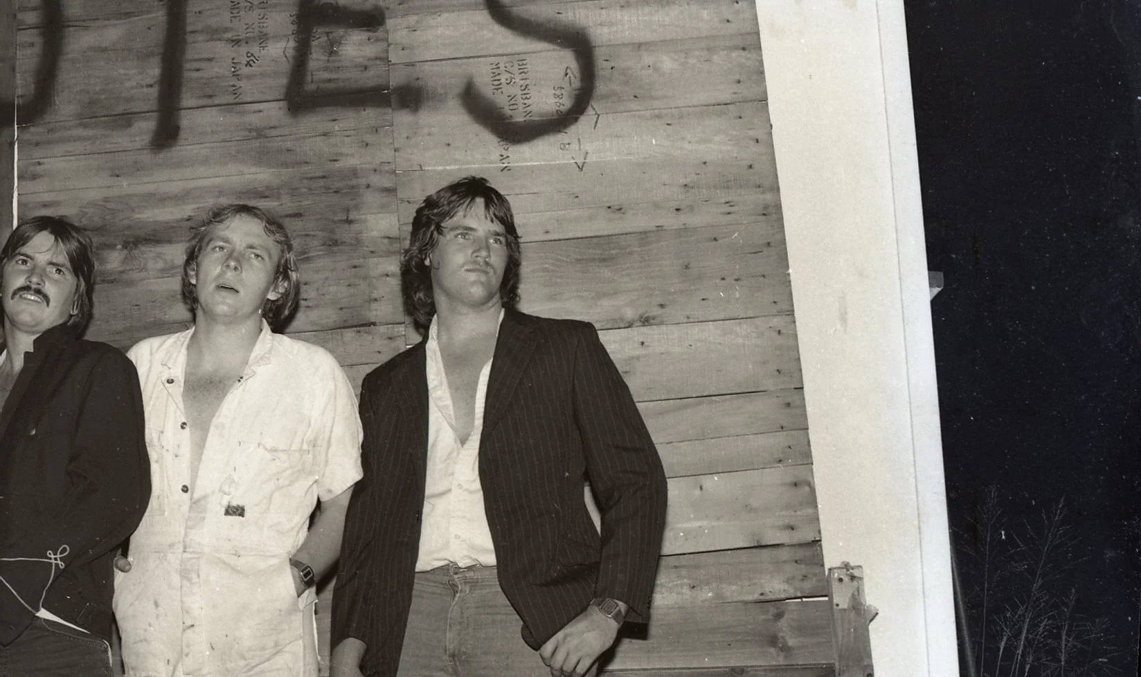 Three men standing in front of a wooden wall at night, one wearing a suit and the other two in casual shirts.