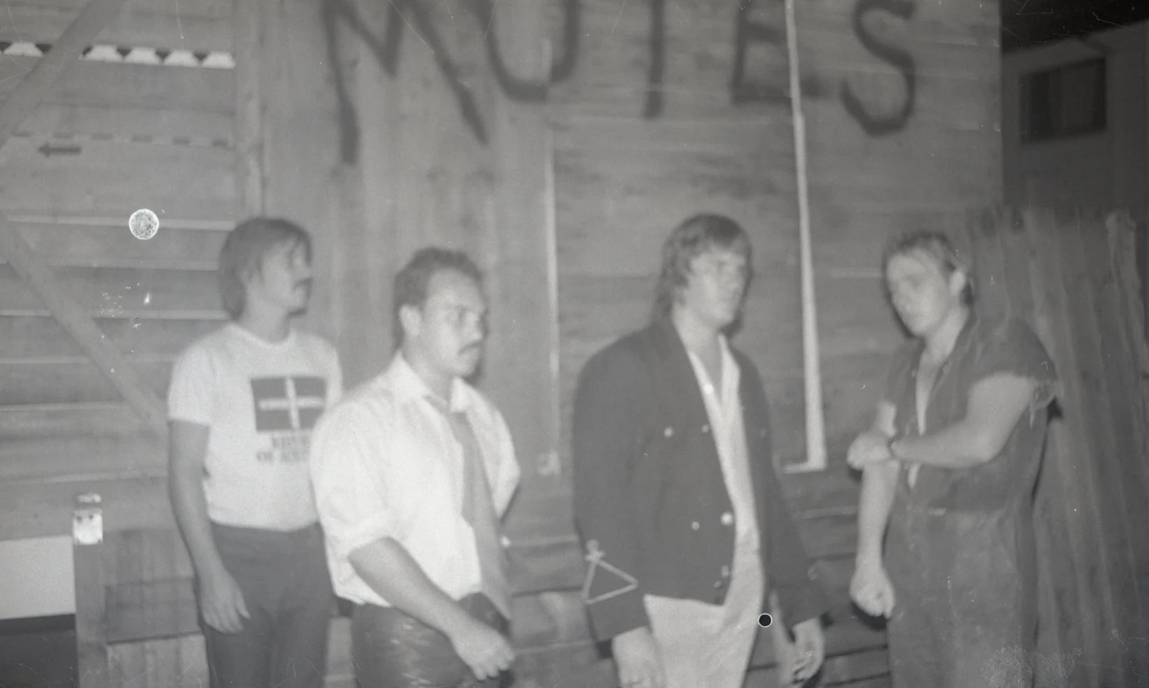 Four men standing indoors against a wooden wall. They are having a conversation. One man is wearing a jacket, two are in shirts, and one is in a T-shirt with a cross and flag design.