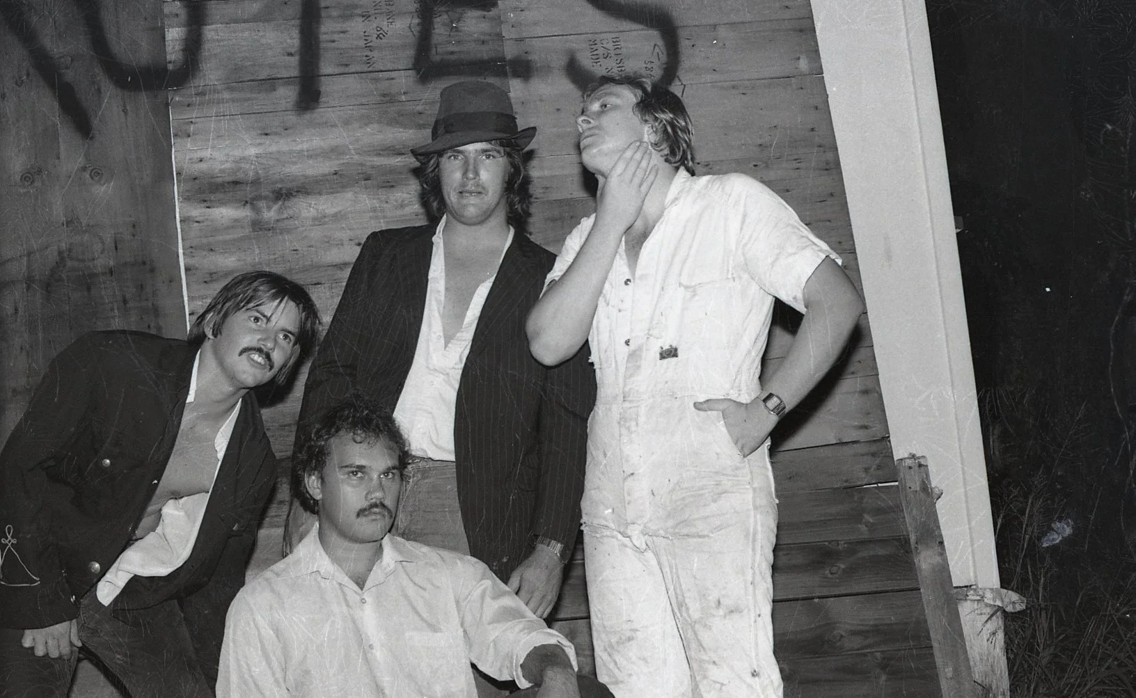 Black and white photo of five men posing together against a wooden wall, with some graffiti or markings on the wall.