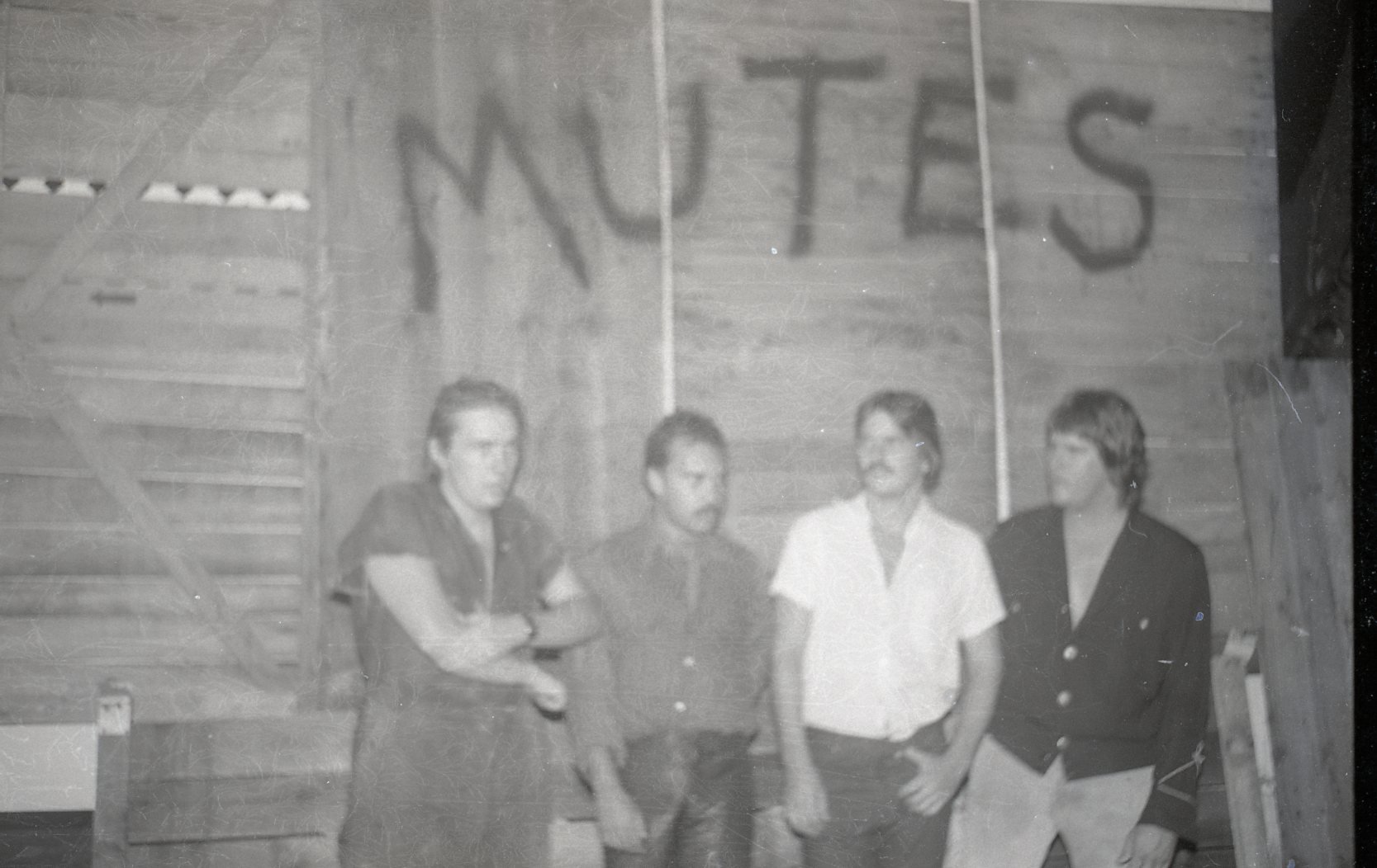 Four people standing in front of a wooden wall with the letters 'NUTS' spray-painted on it.