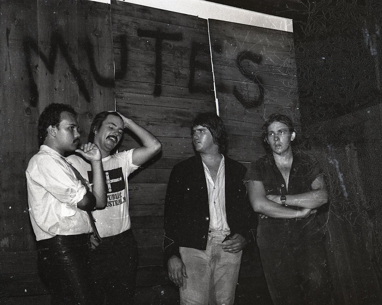 Four young men standing against a wooden wall, engaged in conversation, with a large cross painted on the wall behind them.