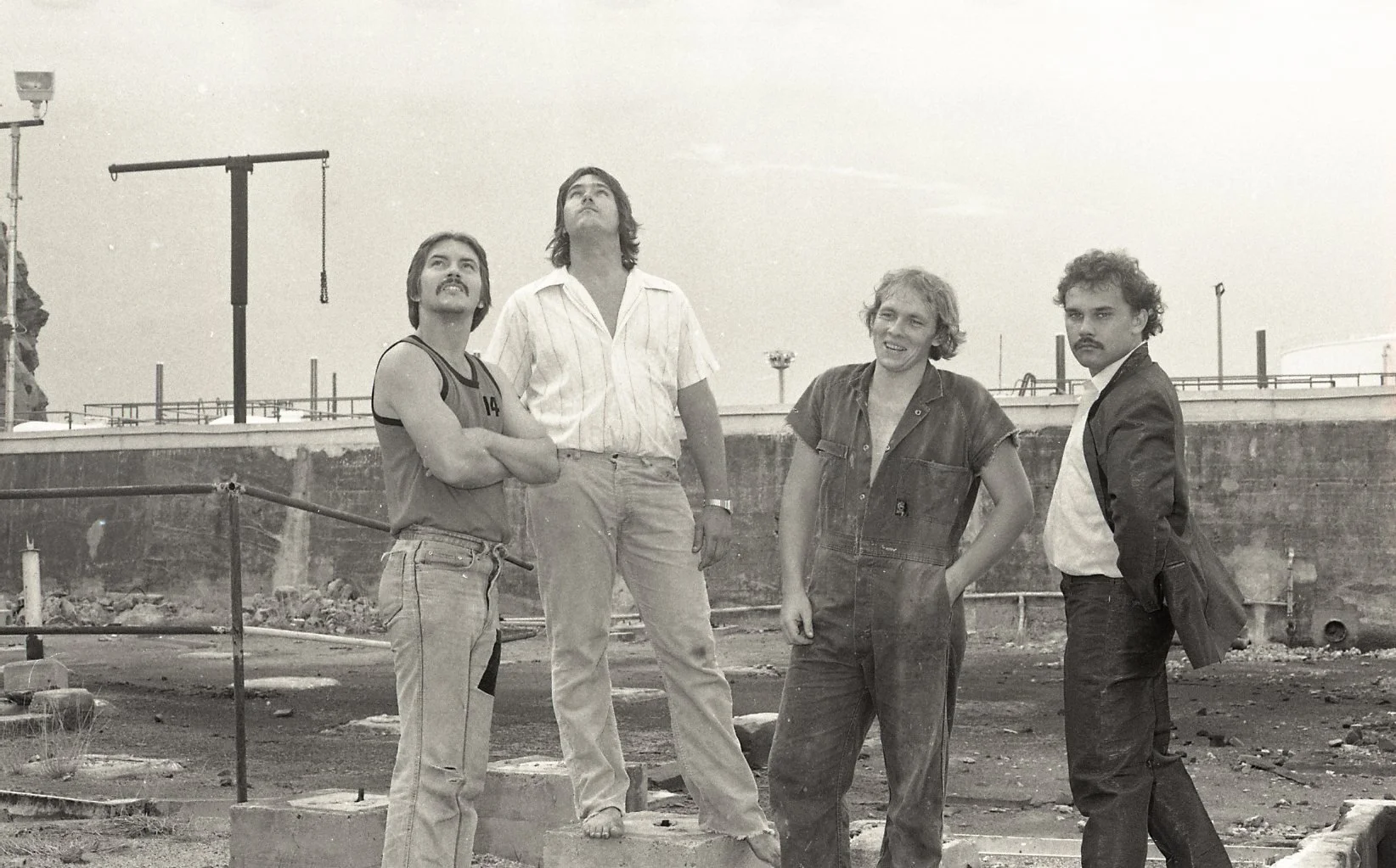 Four men standing outdoors near a construction site with rocks and metal fencing, with a cloudy sky in the background.