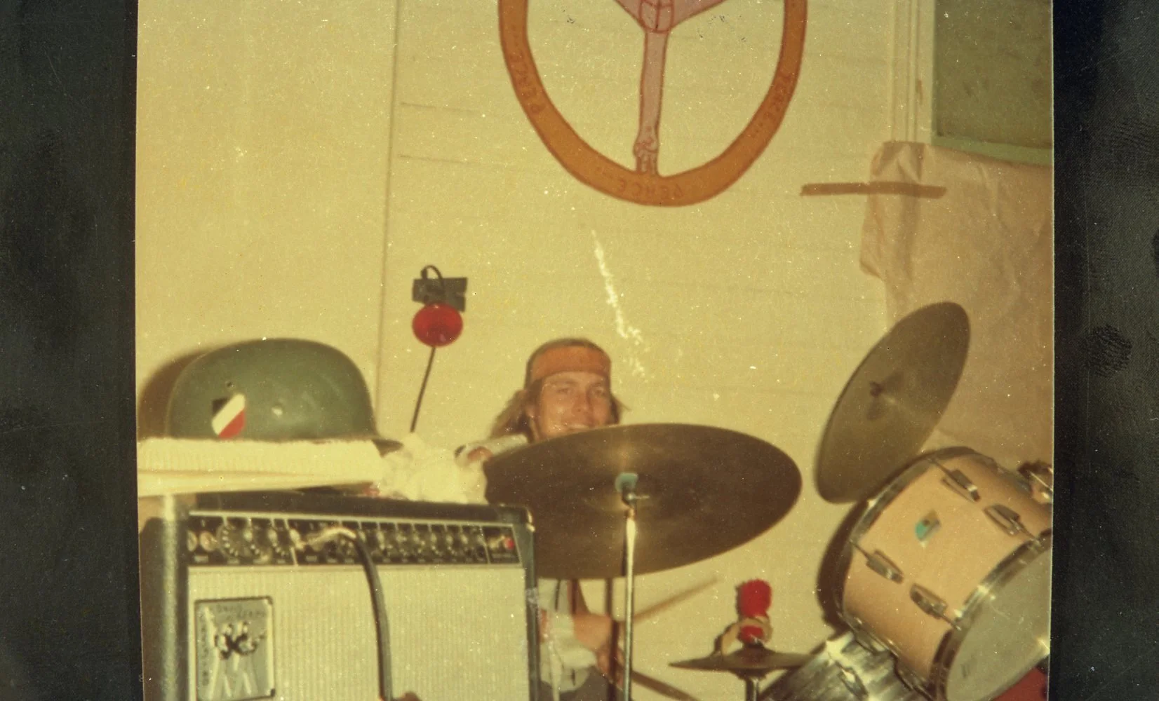 A person playing drums behind musical equipment, with military helmet and mask on a table, and a wall with a martial arts poster.