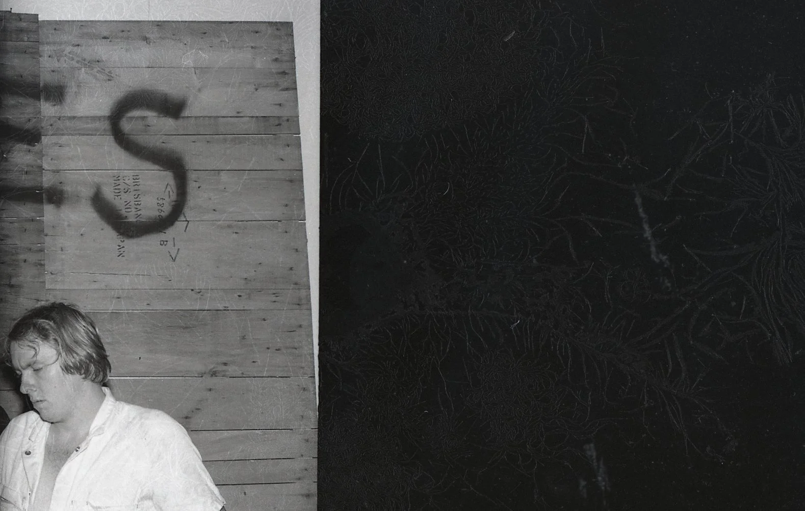 A black and white photo of a young man with short hair, wearing a white shirt, sitting with his eyes closed. The background is a wooden wall, and there is a shadow on the wall that resembles the letter 'S'.