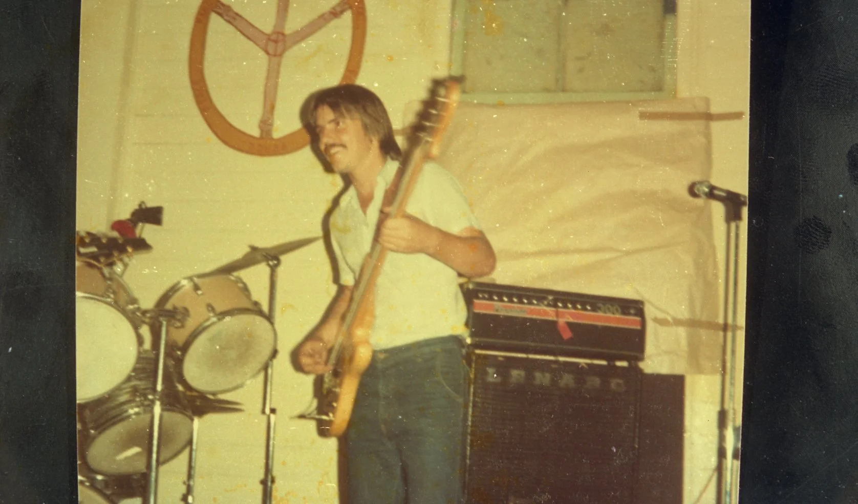 A man playing guitar and singing in a music practice space, with drums, a guitar amplifier, a microphone, and a peace symbol painting on the wall behind him.