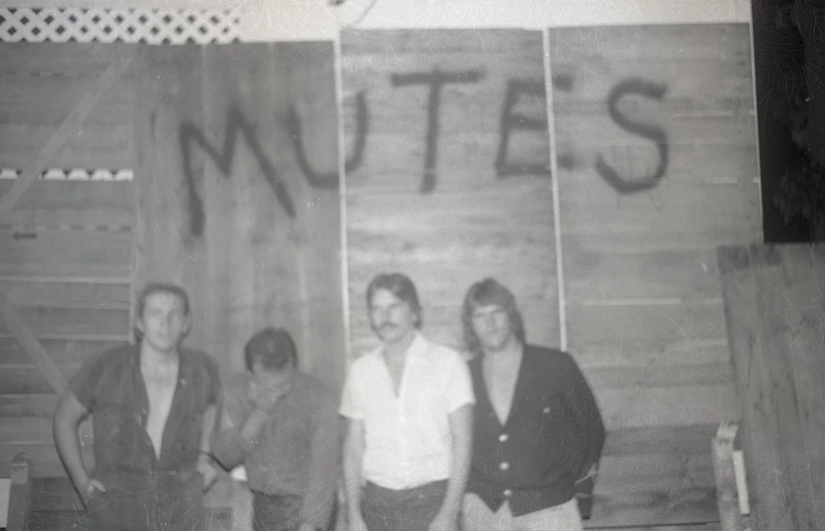 Black and white photo of four people standing in front of a wooden wall with the word 'WOTES' written on it. The individuals appear to be posing for a photo, with one woman and three men, dressed in casual and semi-formal attire.