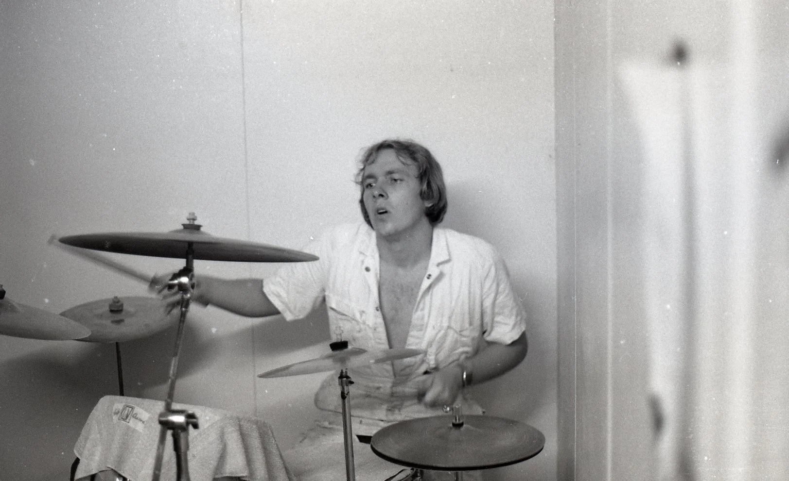 A young man with shoulder-length hair playing a drum set in a small room, wearing a partially unbuttoned white shirt.