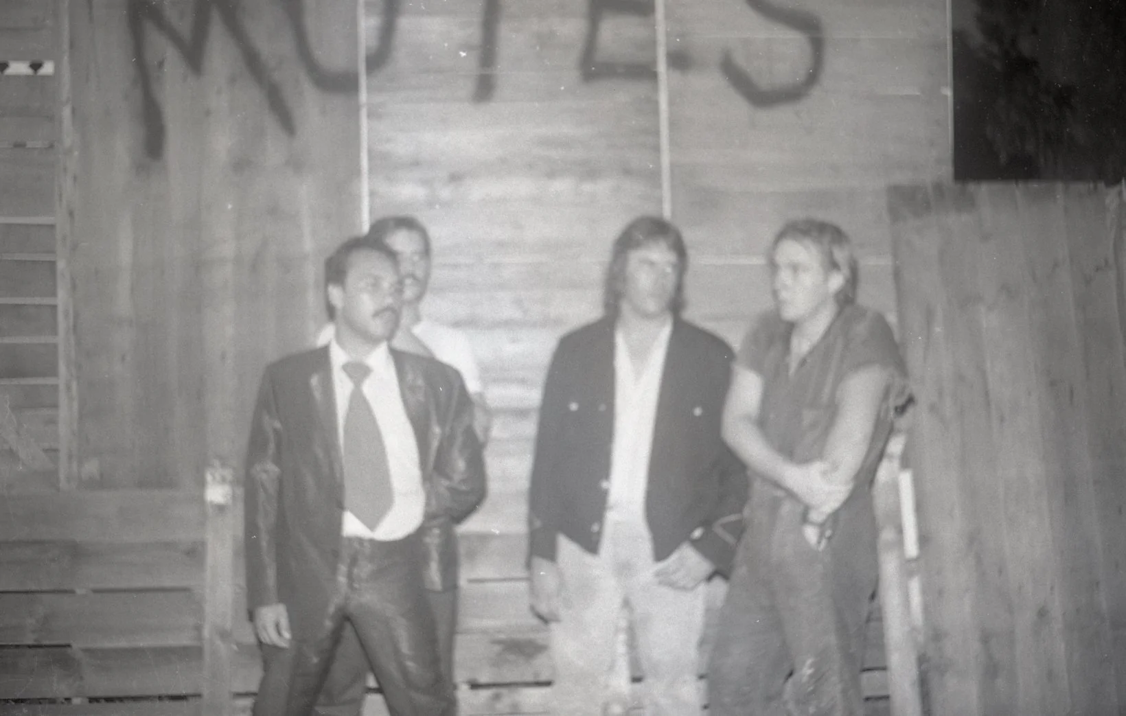Four people standing in a wooden room, dressed in old-fashioned clothing, posing for a photo. The background is wooden panels, and part of a sign is visible at the top.