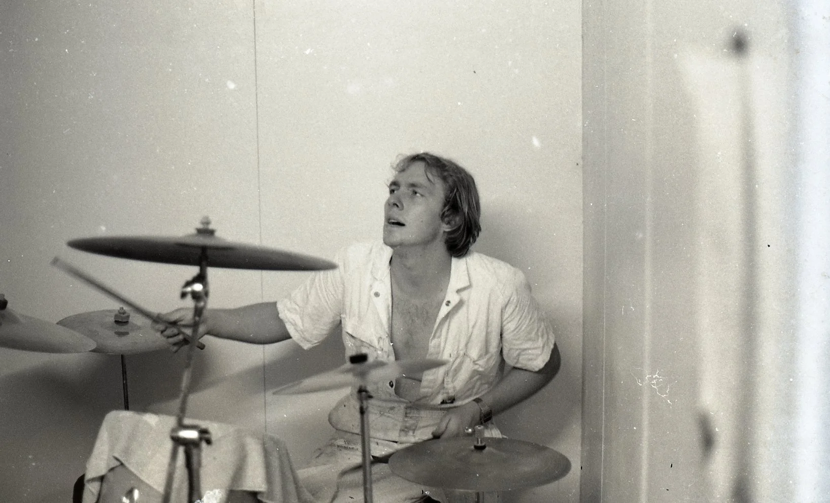 Black and white photo of a man with medium-length hair playing a drum set, holding a drumstick in his right hand, seated against a plain wall.