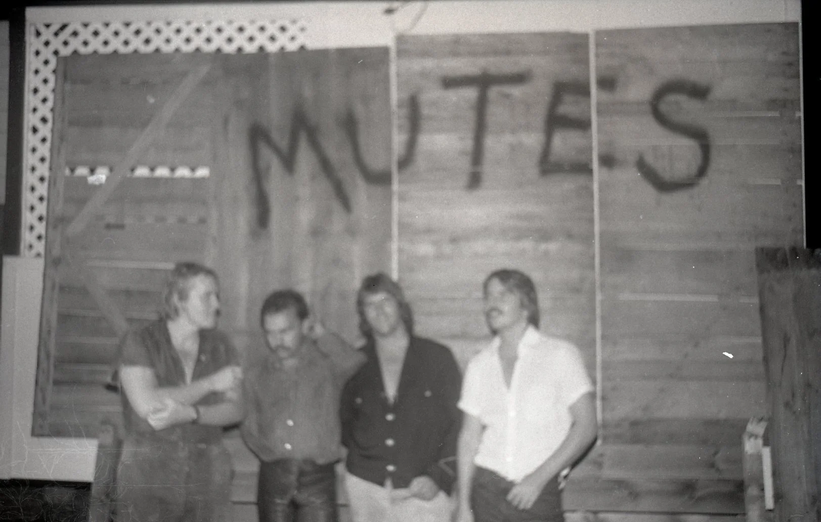Black and white photo of four people standing in front of a wooden wall with a banner that has the word 'MUTAS' written on it. The group includes three women and one man, all engaged in conversation or looking at each other.