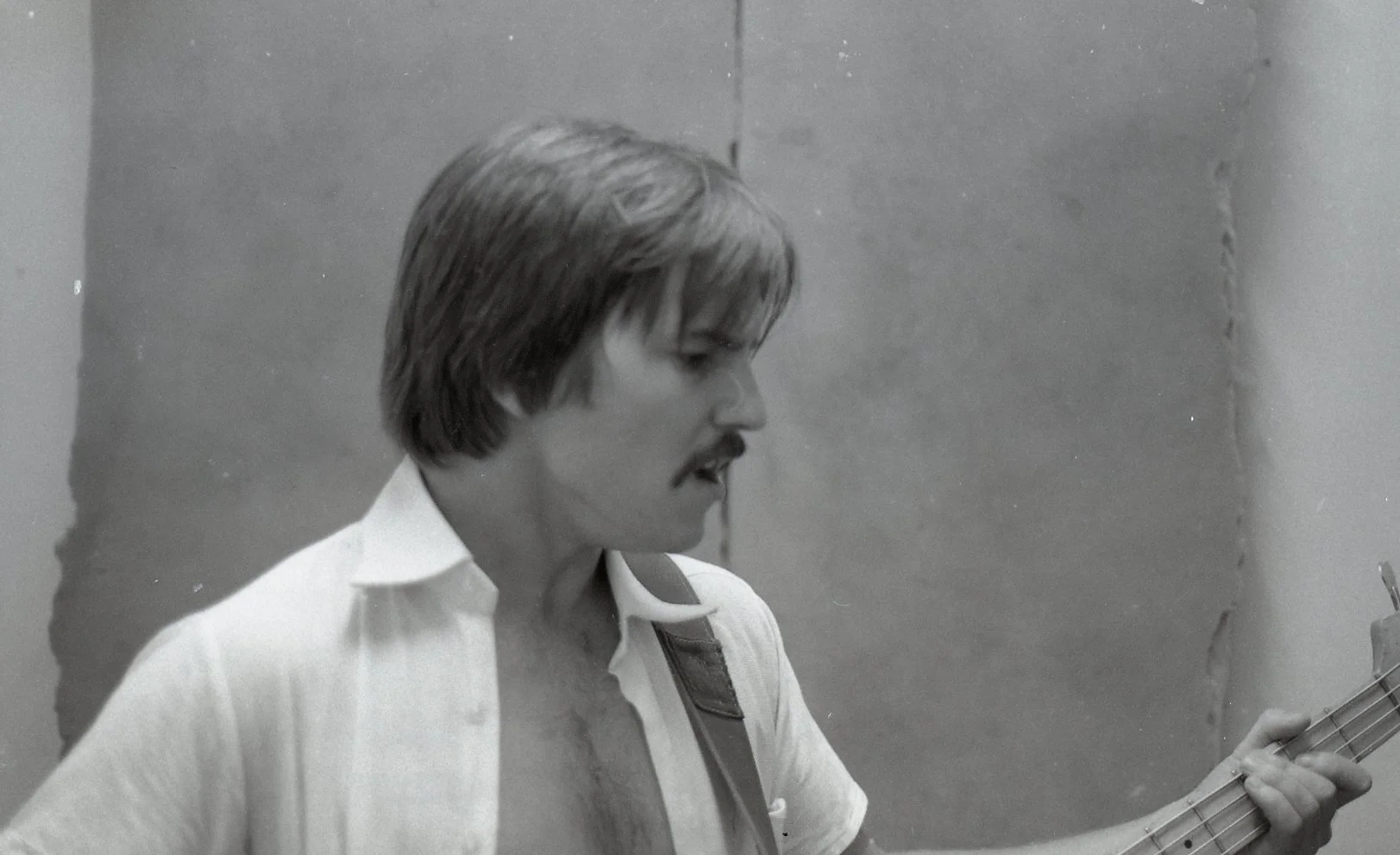 Black and white photo of a man with a mustache, shoulder-length hair, playing an acoustic guitar in a room with plain walls.