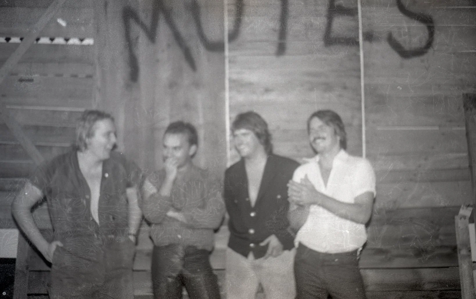 Four women standing together indoors, engaged in a conversation and smiling, with a wooden wall in the background.