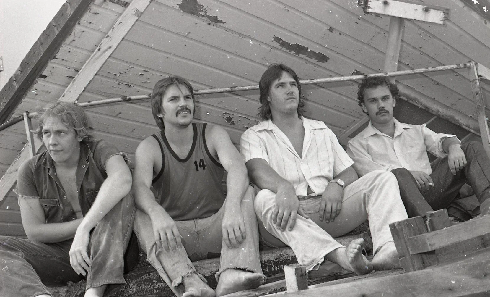 Black and white photo of four men sitting on a weathered wooden surface under an inclined structure, casually dressed, with serious or contemplative expressions.