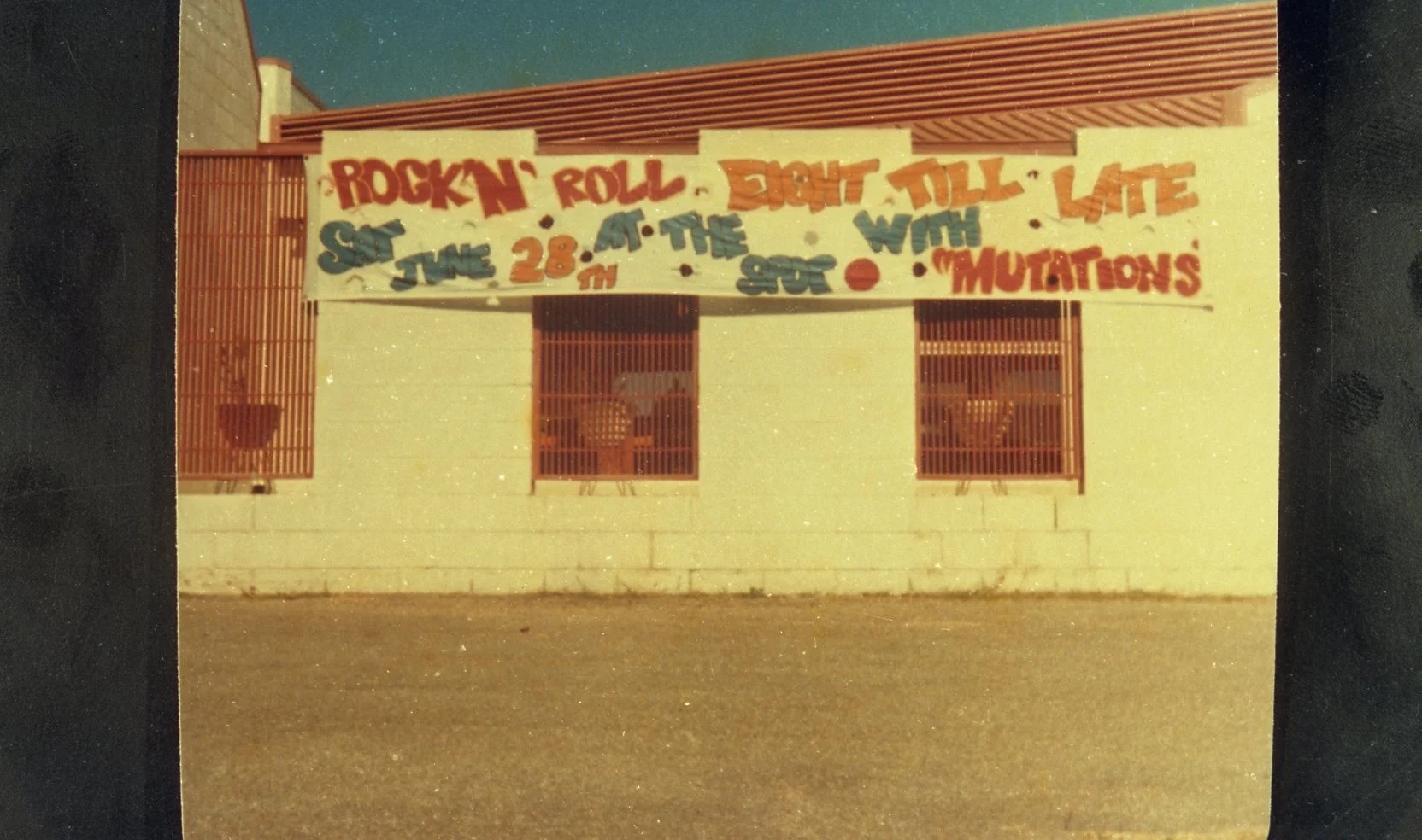 Building with a large colorful banner advertising a rock and roll event on June 28, featuring names such as the Night Tull and the Mutations, with two windows with bars and plants hanging outside.