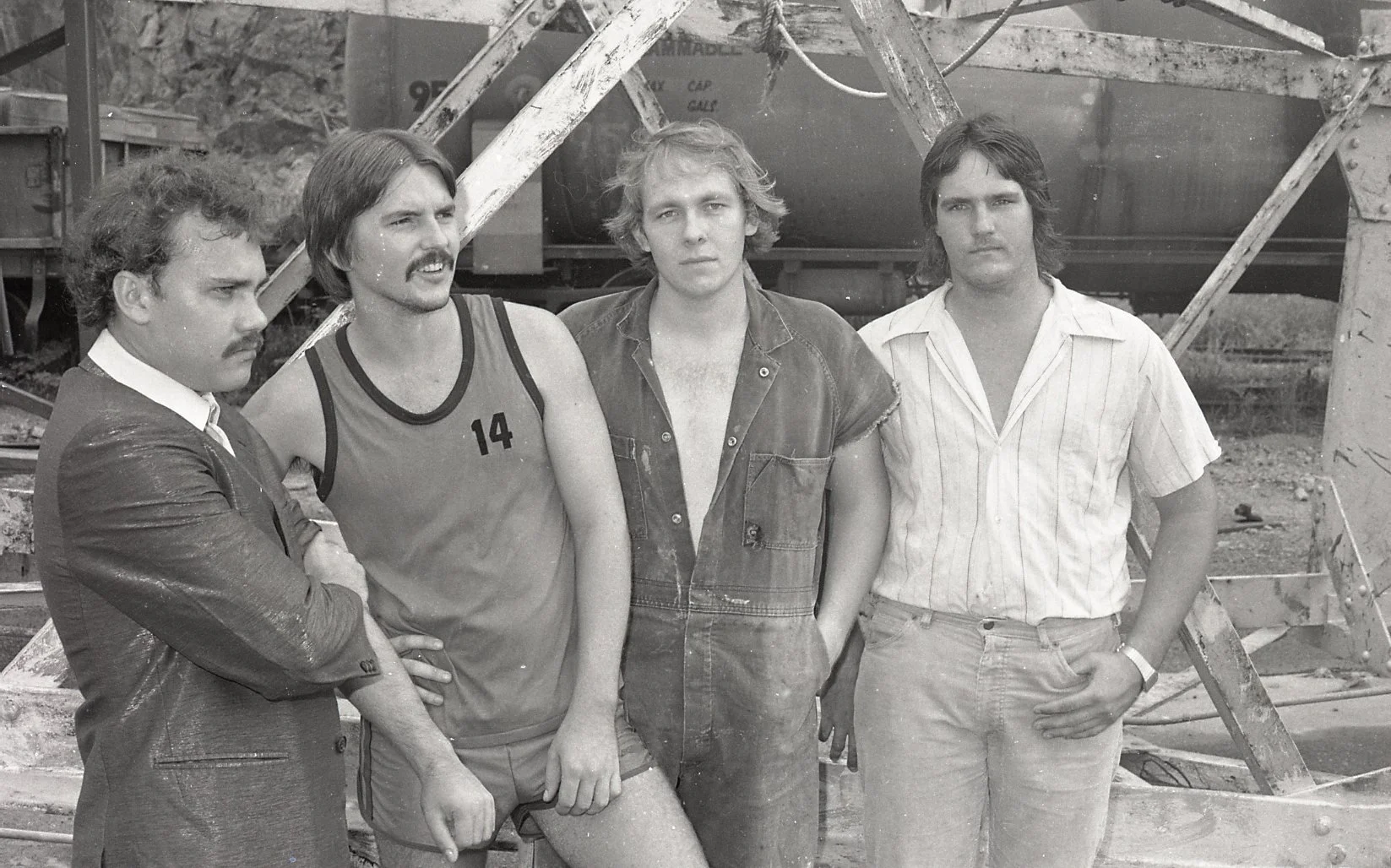 Black and white photo of four men standing outdoors on a construction site, with construction materials and dirt in the background. One man is dressed in a suit, the second man is wearing a sports jersey with the number 14, the third man is wearing a