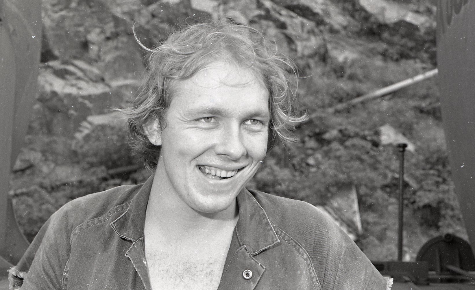 Black and white photo of a young man with wavy hair smiling, wearing a denim shirt, with a rocky background.