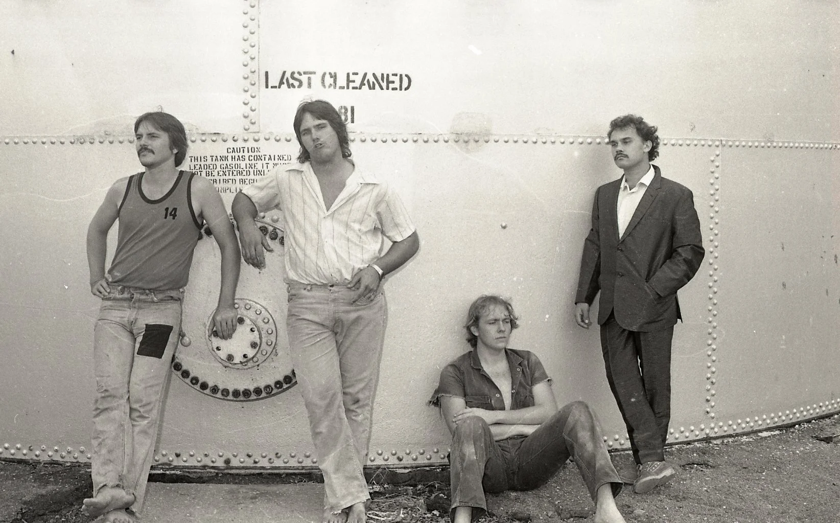 Four young people with casual and formal attire posing against a large industrial tank. One person is sitting on the ground with crossed arms, the others are standing or leaning, looking in different directions. The tank has a sign that says 'LAST CL