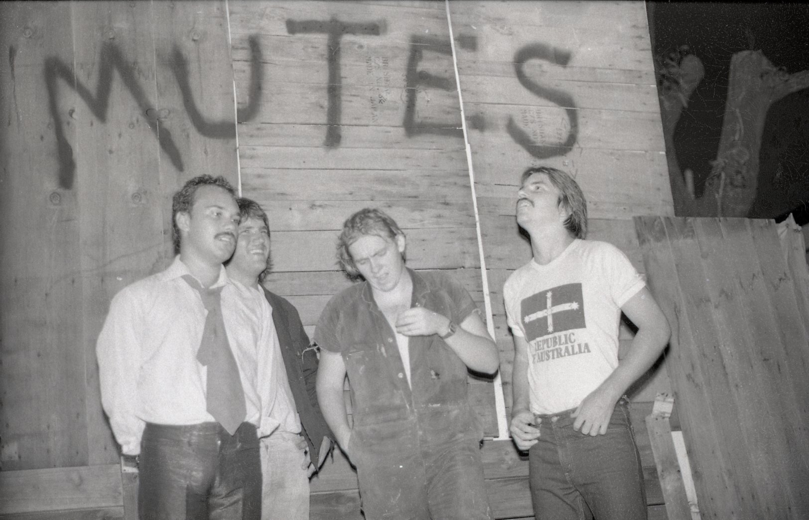 Four young men standing against a wooden wall, with shadows of letters spelling 'WET'S' cast on the wall behind them. One man wears a tie and a white shirt, another wears a dark jacket, the third has curly hair and wears a denim vest, and the fourth 