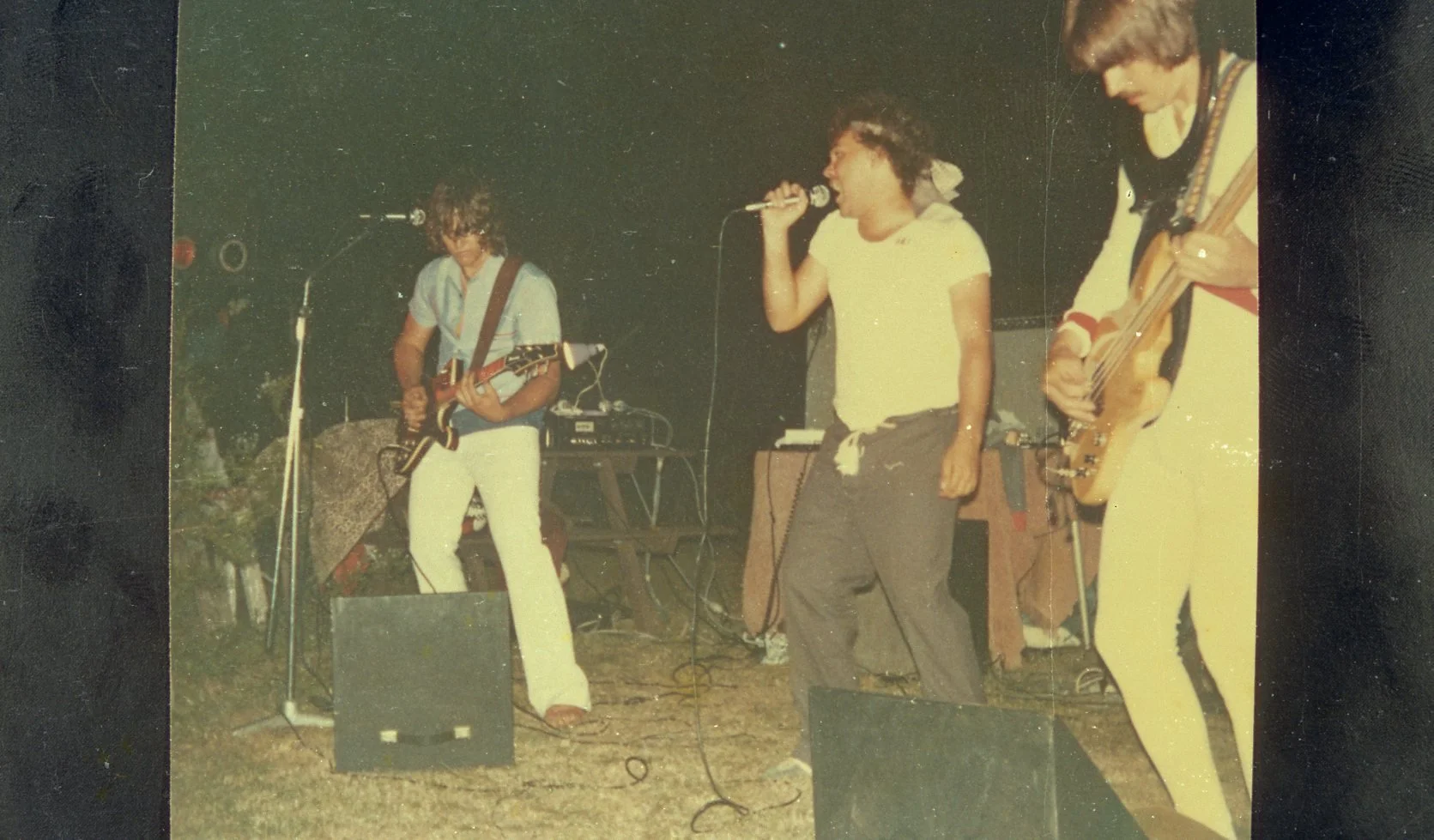 Three young musicians performing outdoors at night with guitars and microphone.