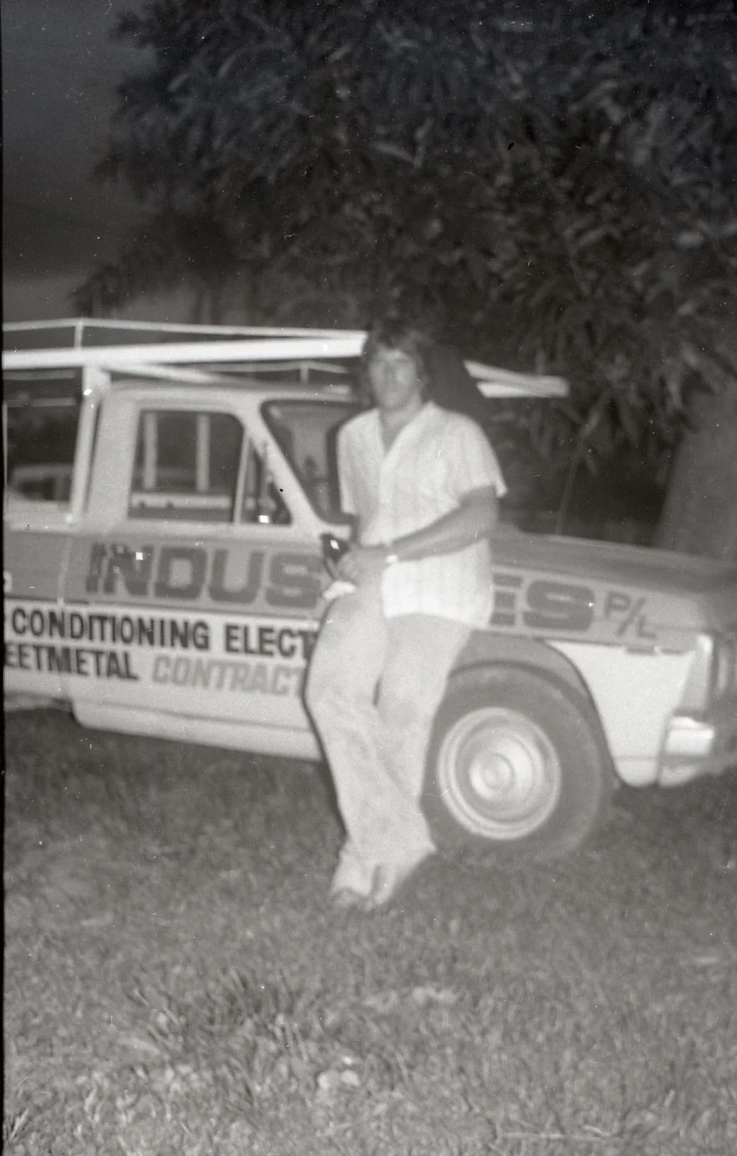 A young woman sitting on the side of a pickup truck outdoors at night, with a large tree behind her. The truck advertises a heating and air conditioning business.