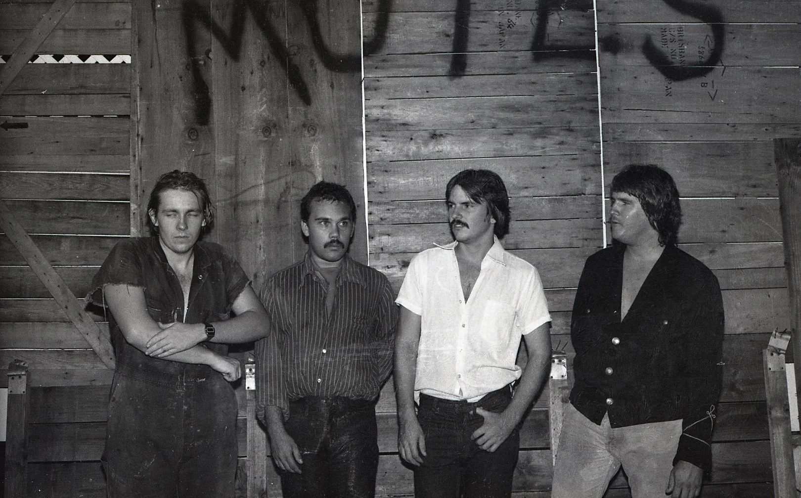 Black and white photo of four men standing against a wooden wall. The first man on the left has long hair and is wearing a short-sleeve shirt with his arms crossed. The second man has short hair, a mustache, and is wearing a striped shirt. The third 