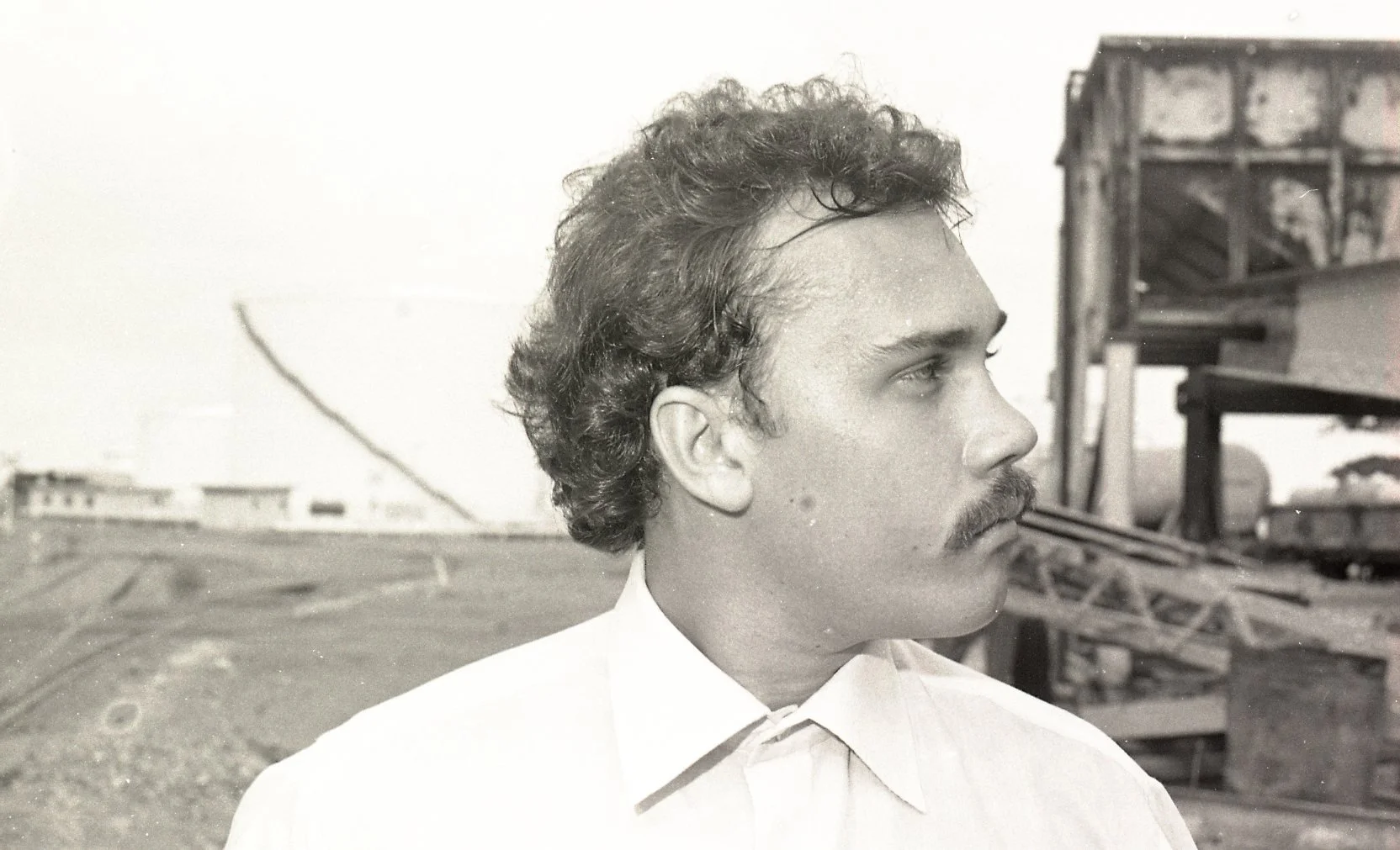 Black and white photograph of a man with curly hair and a mustache, wearing a collared shirt, standing outdoors on a rooftop with industrial structures in the background.