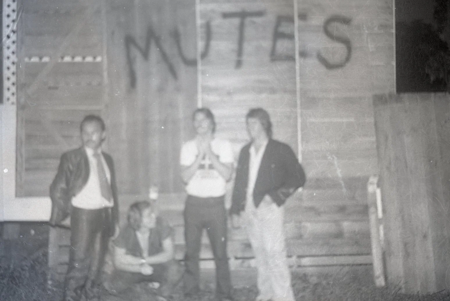 Four people standing in front of a wooden wall with the word 'NUTS' written above them. Two women are standing, one with glasses and one with glasses, and two men are standing, one with hands in pockets and the other with hands on hips. There is a wo