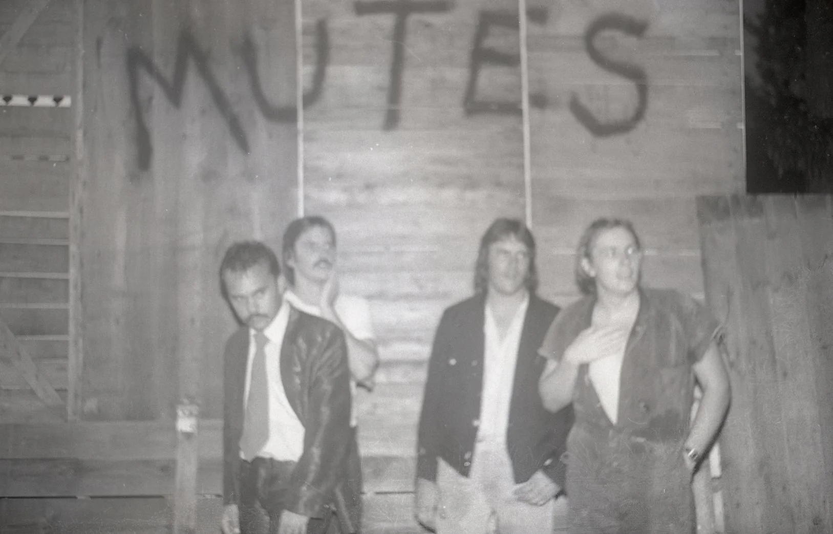 Black-and-white photo of four men standing against a wooden wall with the word 'METEORS' spray-painted above them. The men are dressed in casual and semi-formal clothing, with some wearing jackets. They appear to be posing for the photo.