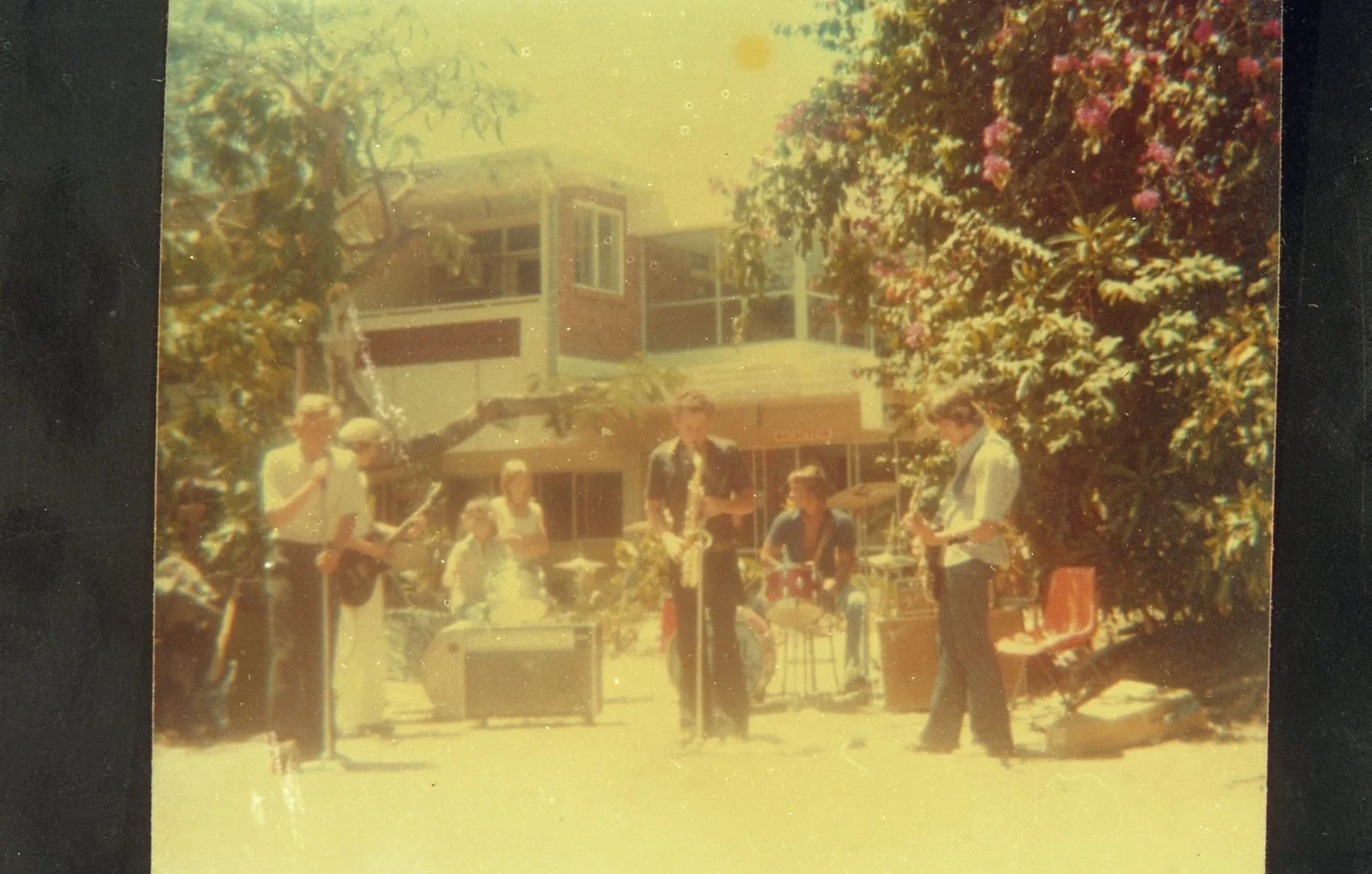 A vintage photo of a small outdoor musical band performing with a house, trees, and pink flowers in the background on a sunny day.