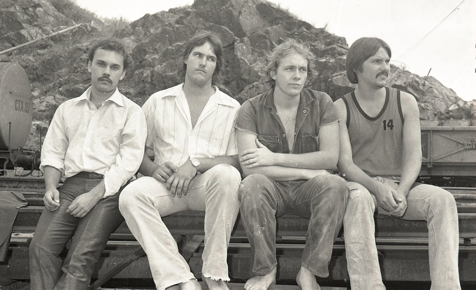 Black and white photo of four men sitting on a train flatbed or platform with rocky terrain in the background. The men are dressed casually, with one wearing a sleeveless shirt with the number 14, and have various hairstyles and facial hair.