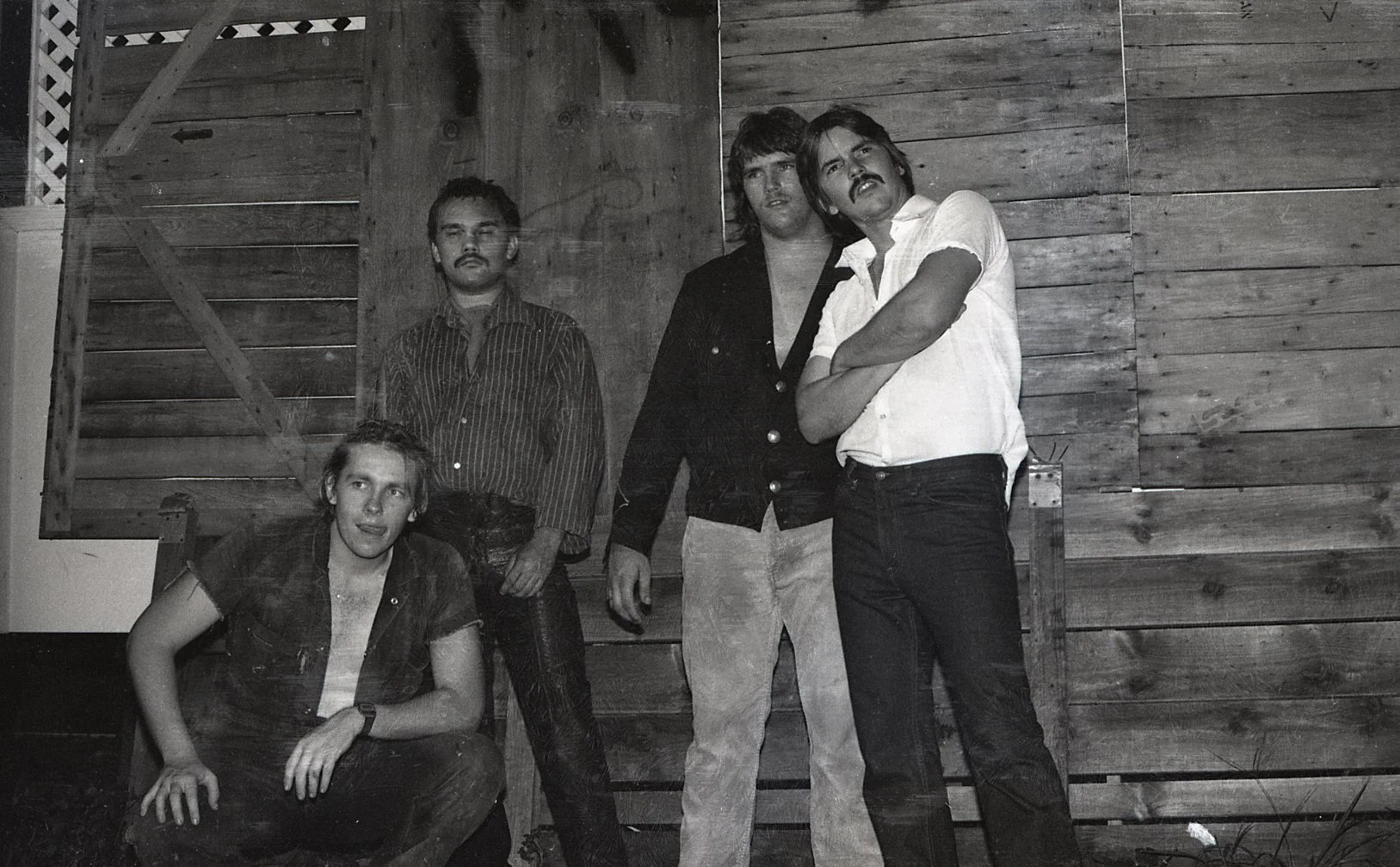 Five men in front of a wooden wall, some standing and one sitting; black and white photograph.