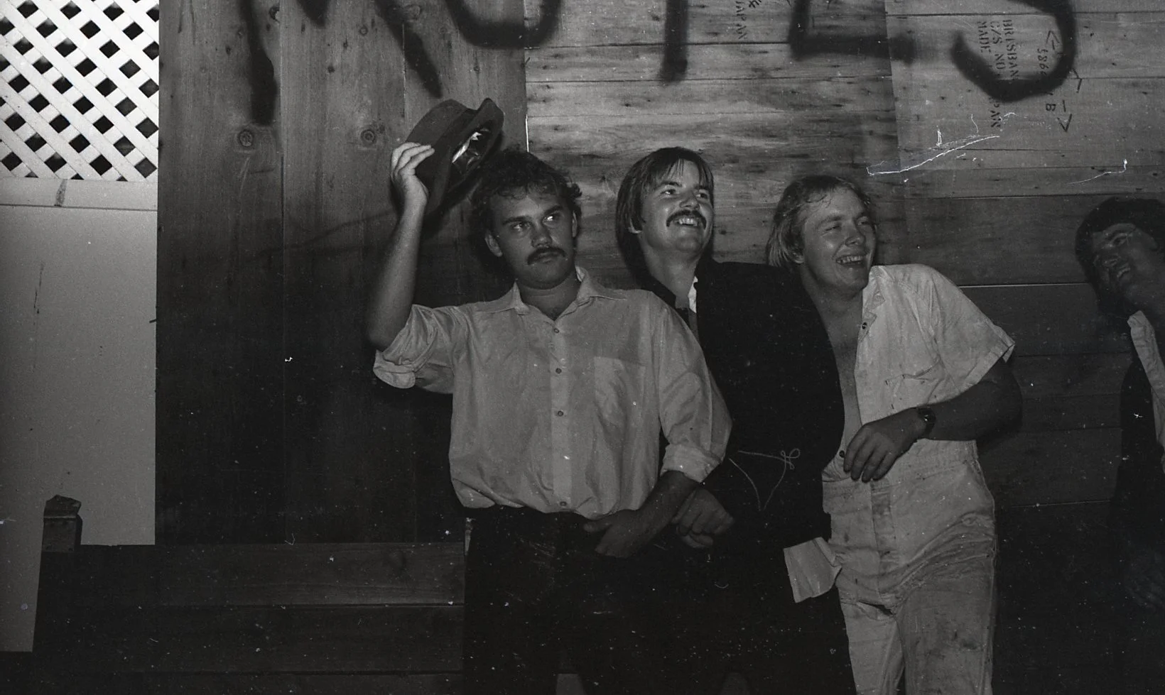 Four men standing against a wooden wall, laughing, one man holding a hat in the air. Black and white photograph, casual setting.