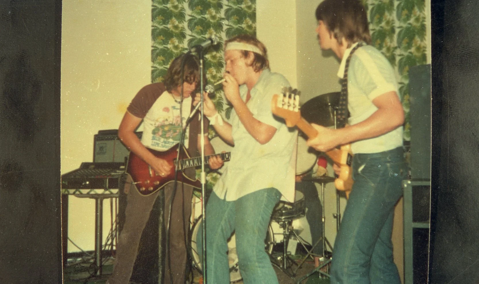 A band of young musicians performing with guitars and a drum set in a room with patterned curtains.