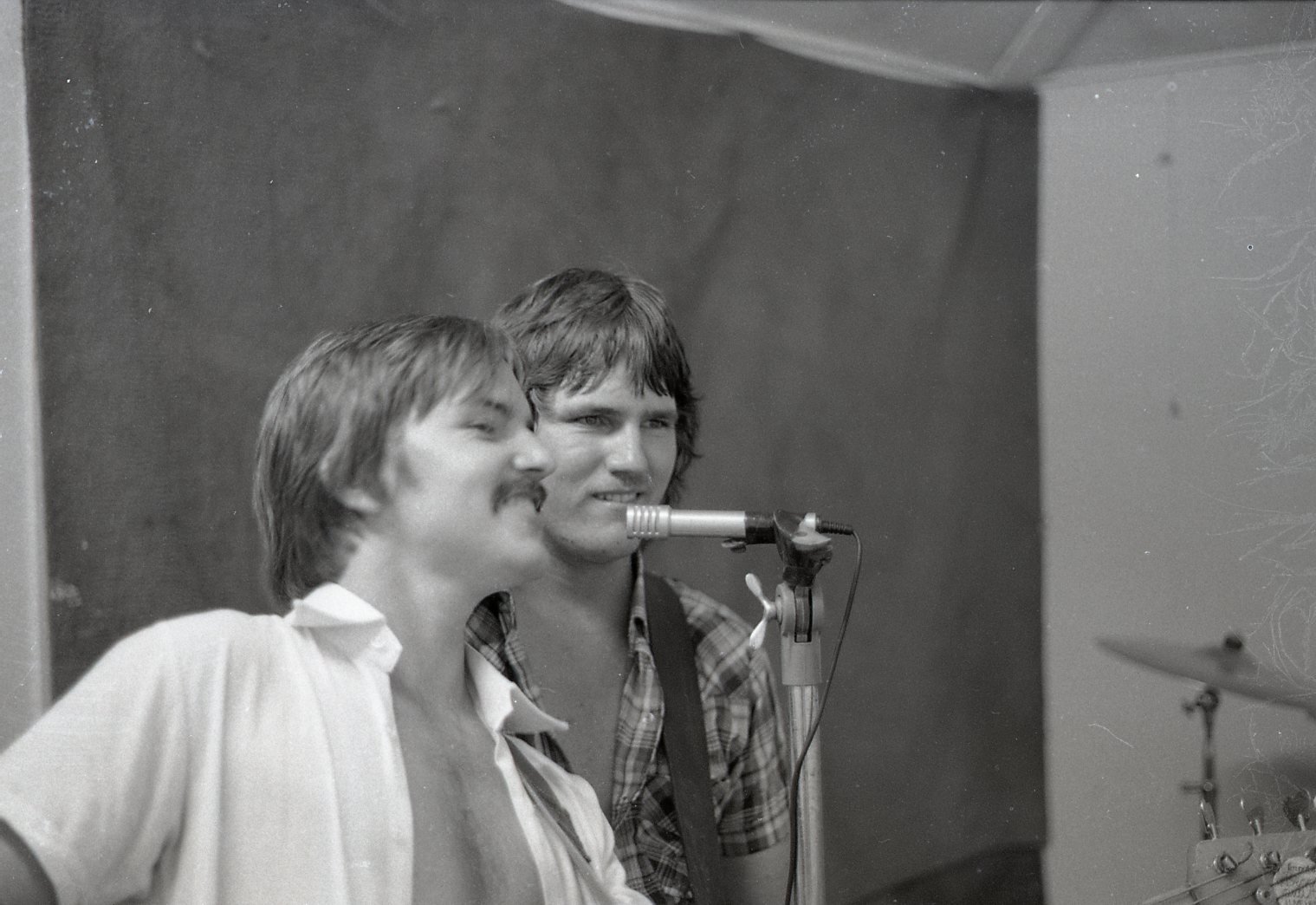 Two young men singing into a microphone in a recording studio, one with a mustache, the other with ash blonde hair, a drum set partially visible on the right.