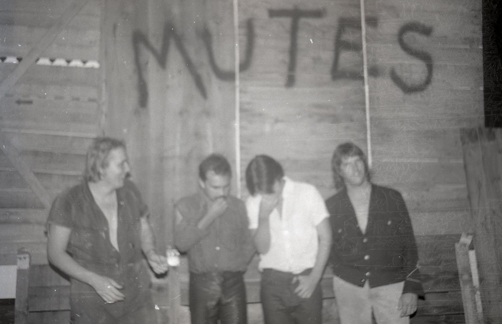 Four people standing together inside a wooden building, with one person holding a drink and two looking down or at each other, and a large banner with the word 'NUTS' in the background
