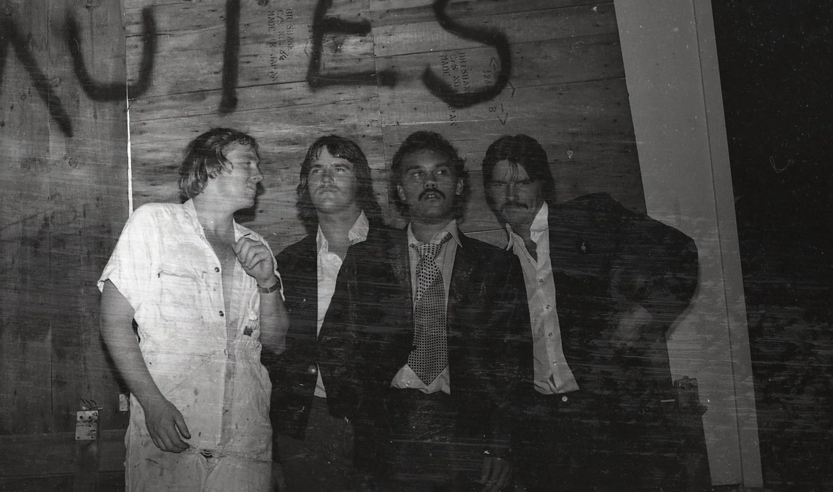 Black and white photo of four men standing against a wooden wall, wearing formal and casual attire. One man is dressed casually in a white shirt and white pants, while the others are in suits and ties.