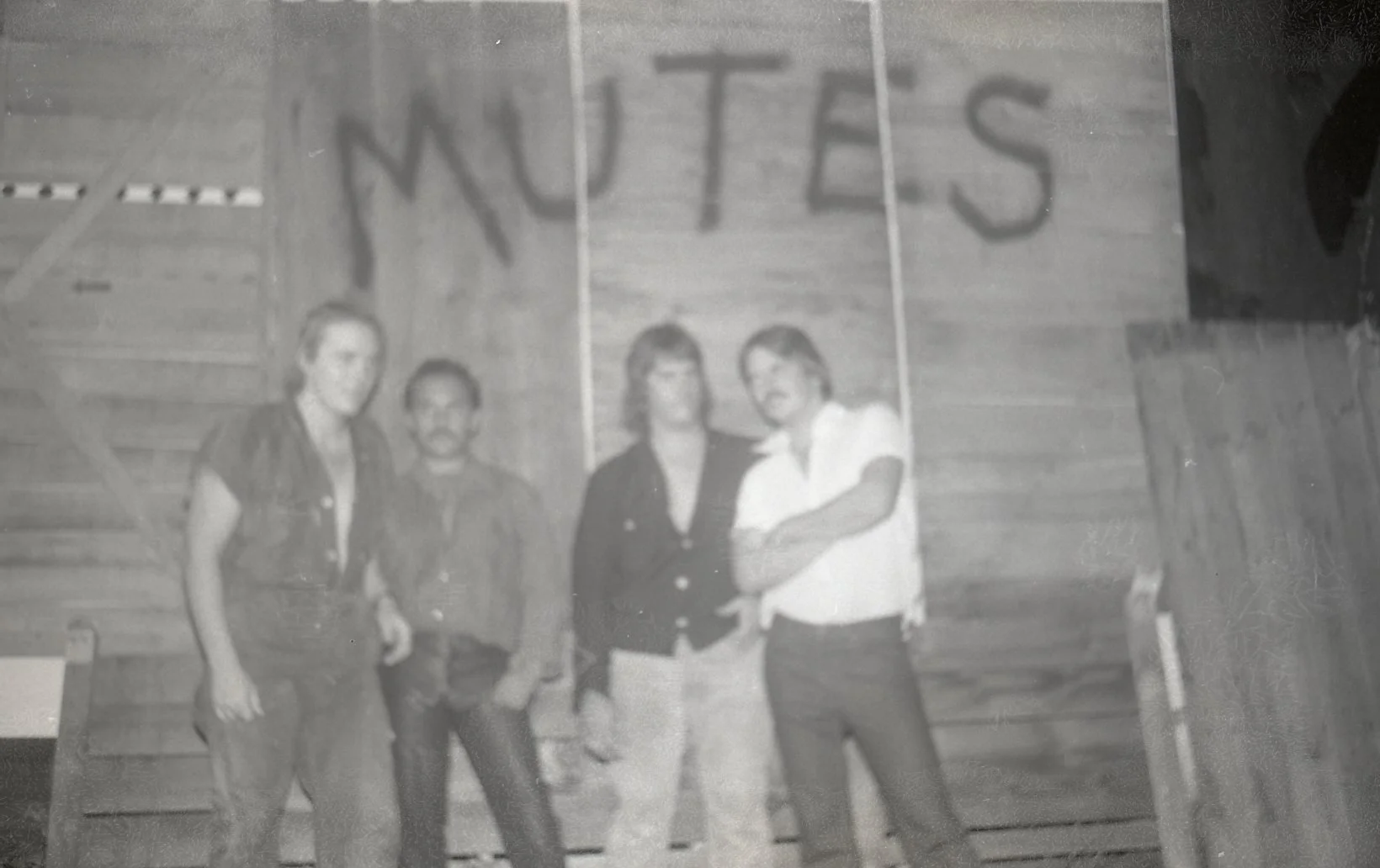 Four people standing in front of a wooden wall with a sign that says "NUTES". They appear to be posing for a photo, with some smiling and some with neutral expressions.
