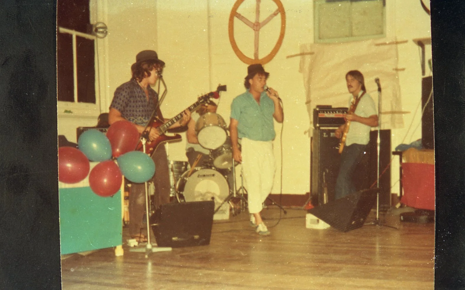 A band performing on a stage decorated with balloons and a peace sign. There are four band members: a woman singing into a microphone, a guitarist, a drummer, and a bassist. The setting appears to be a small indoor event or party.