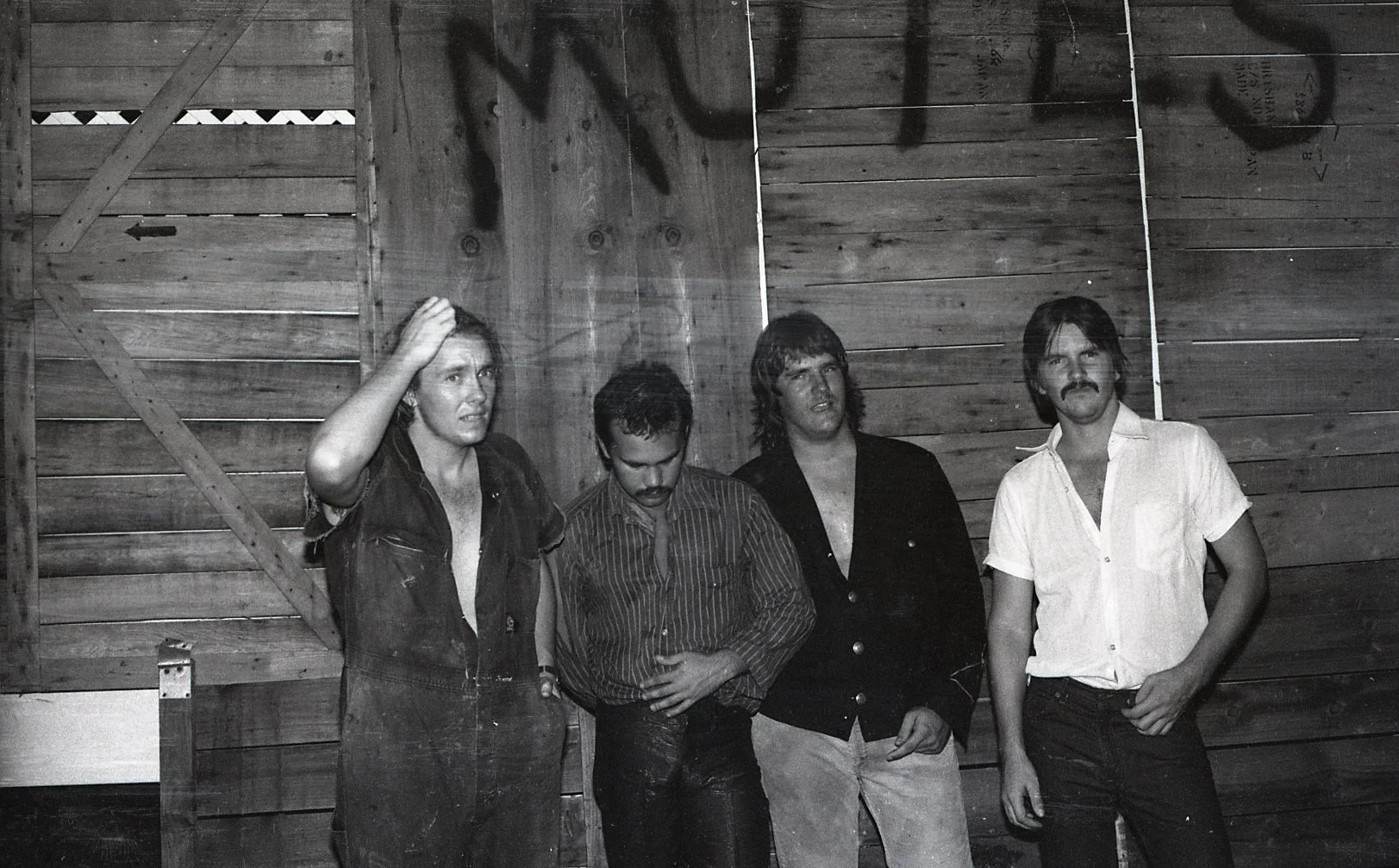 Black and white photo of four men standing against a wooden wall, with some graffiti. The men are casually dressed, some with hand in pockets, one scratching his head, and one with a mustache.
